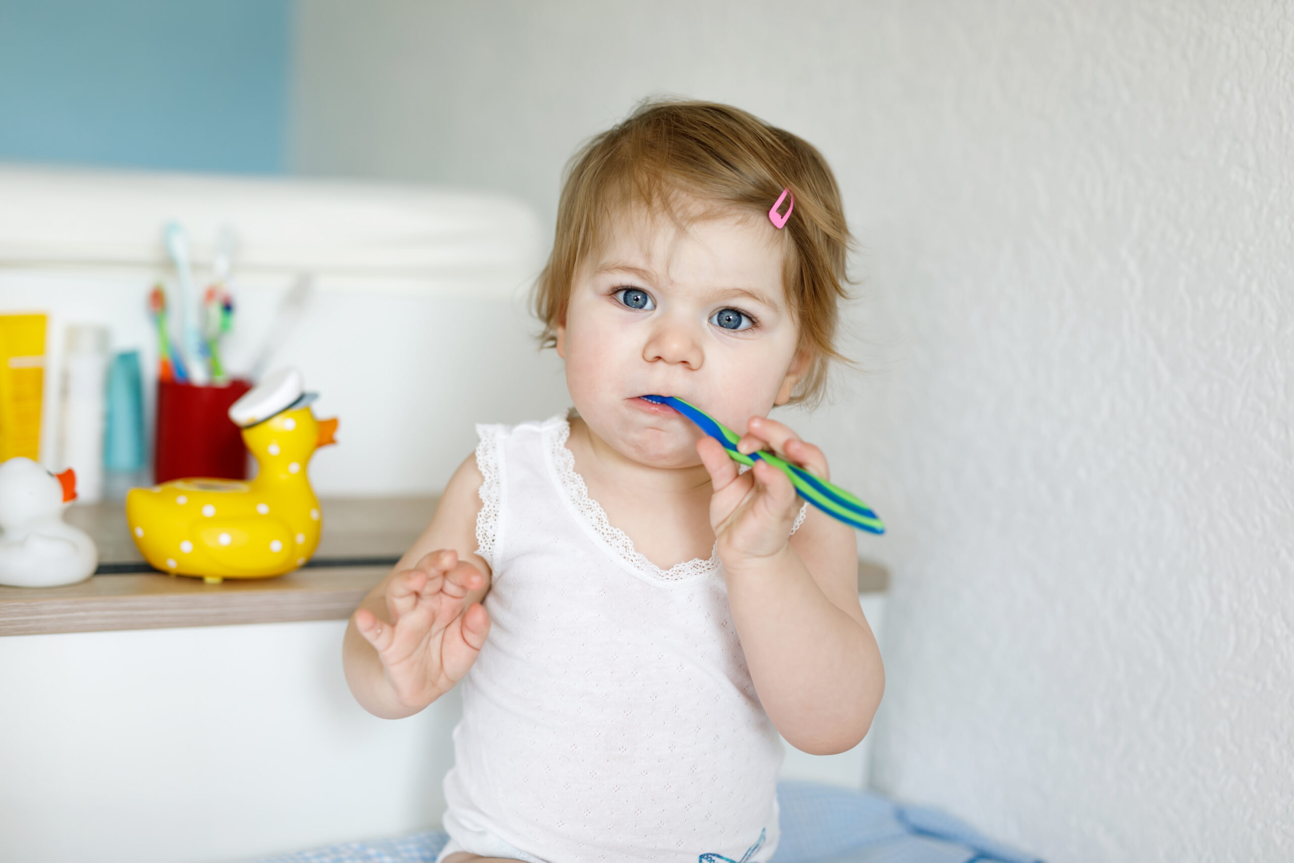 Toddler with blue eyes practices brushing teeth while sitting near bathroom items, holding a colorful toothbrush and looking toward the camera.