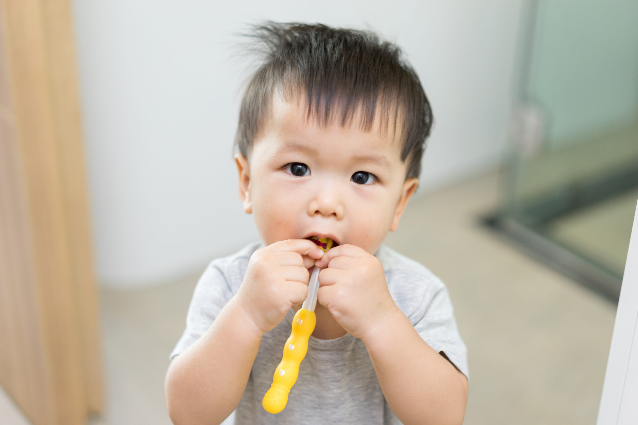 Young child holding a yellow toothbrush practices brushing teeth, standing indoors and looking forward with a focused expression.